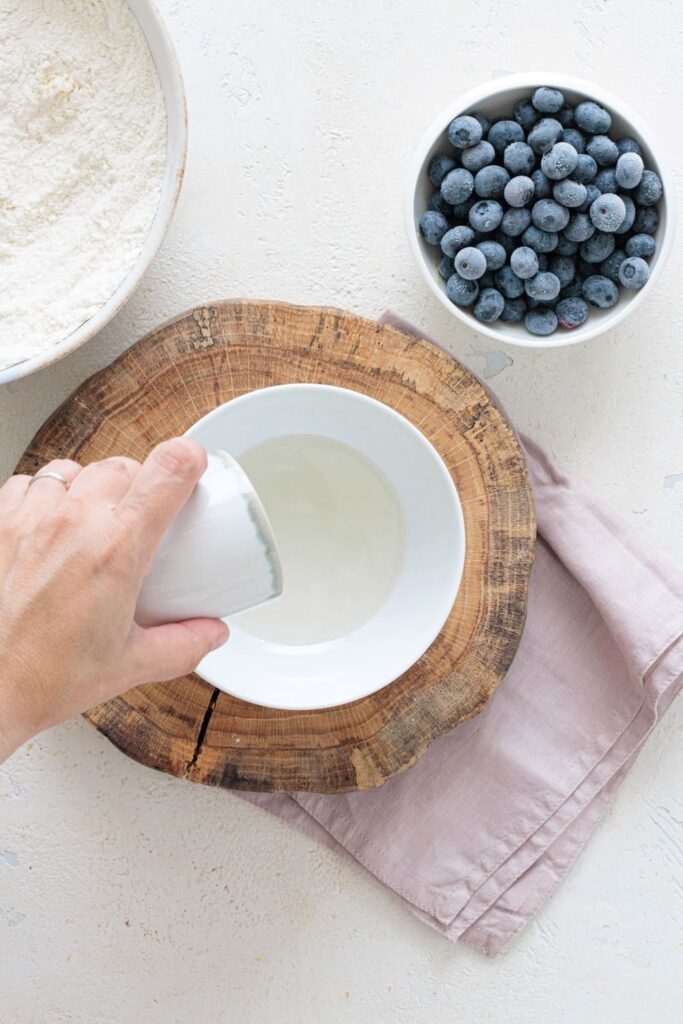 Pouring oil into a mixing bowl with flour and blueberries on the side