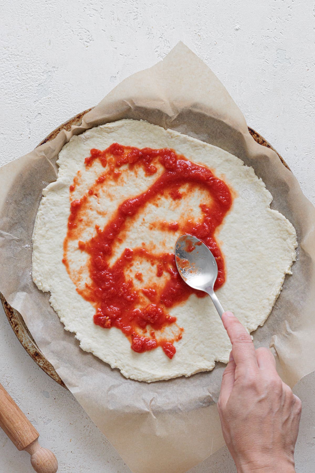 Spreading tomato sauce over Greek yogurt pizza dough base with a spoon.
