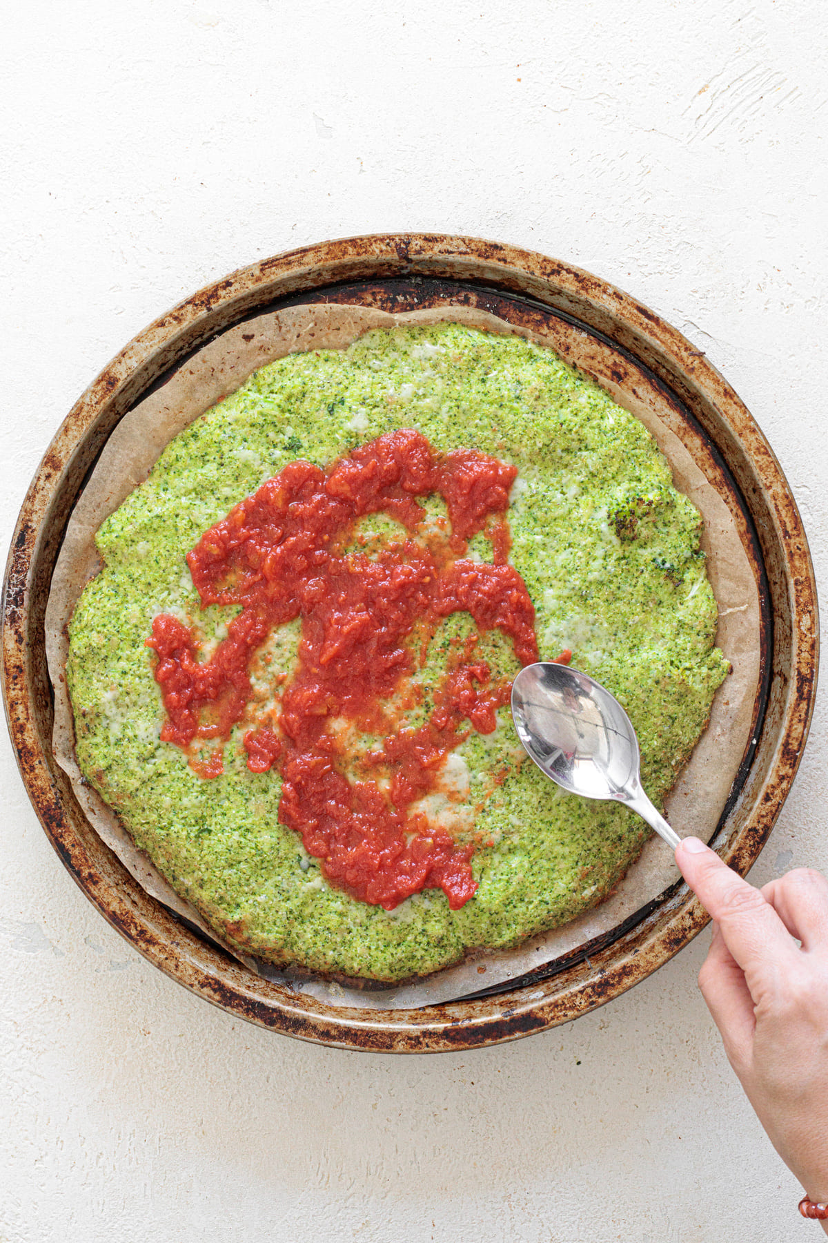 Spreading tomato sauce over the baked broccoli crust with a spoon