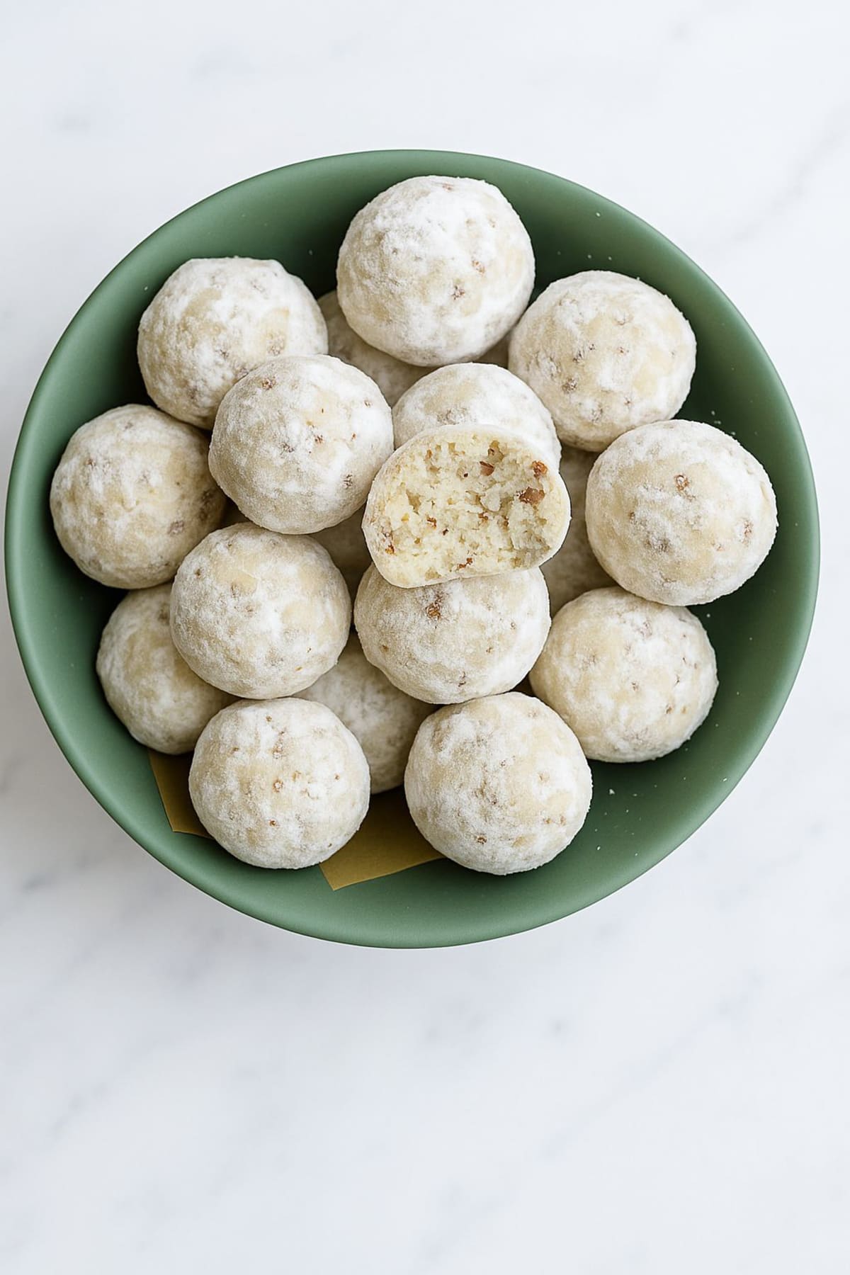 Bowl of Snowball Cookies coated in powdered sugar with one cookie showing its pecan center