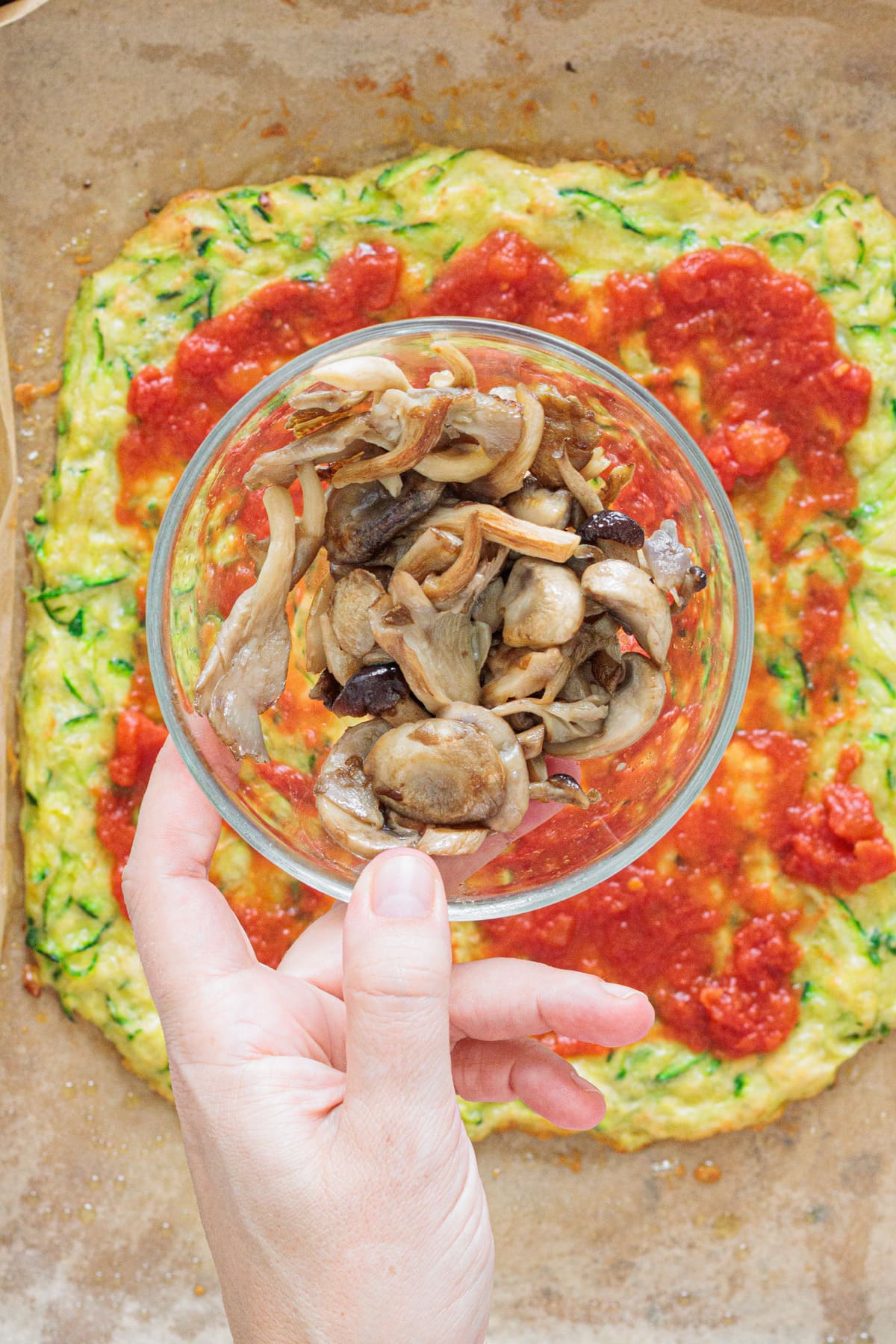 Hand holding a bowl of cooked mushrooms over zucchini crust with tomato sauce